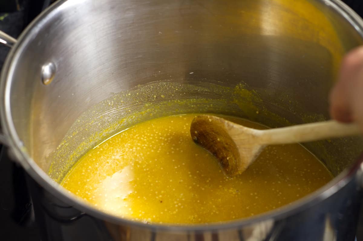 Various spices, vinegar, and sugar being mixed together in a large stainless steel pot with a wooden spoon.