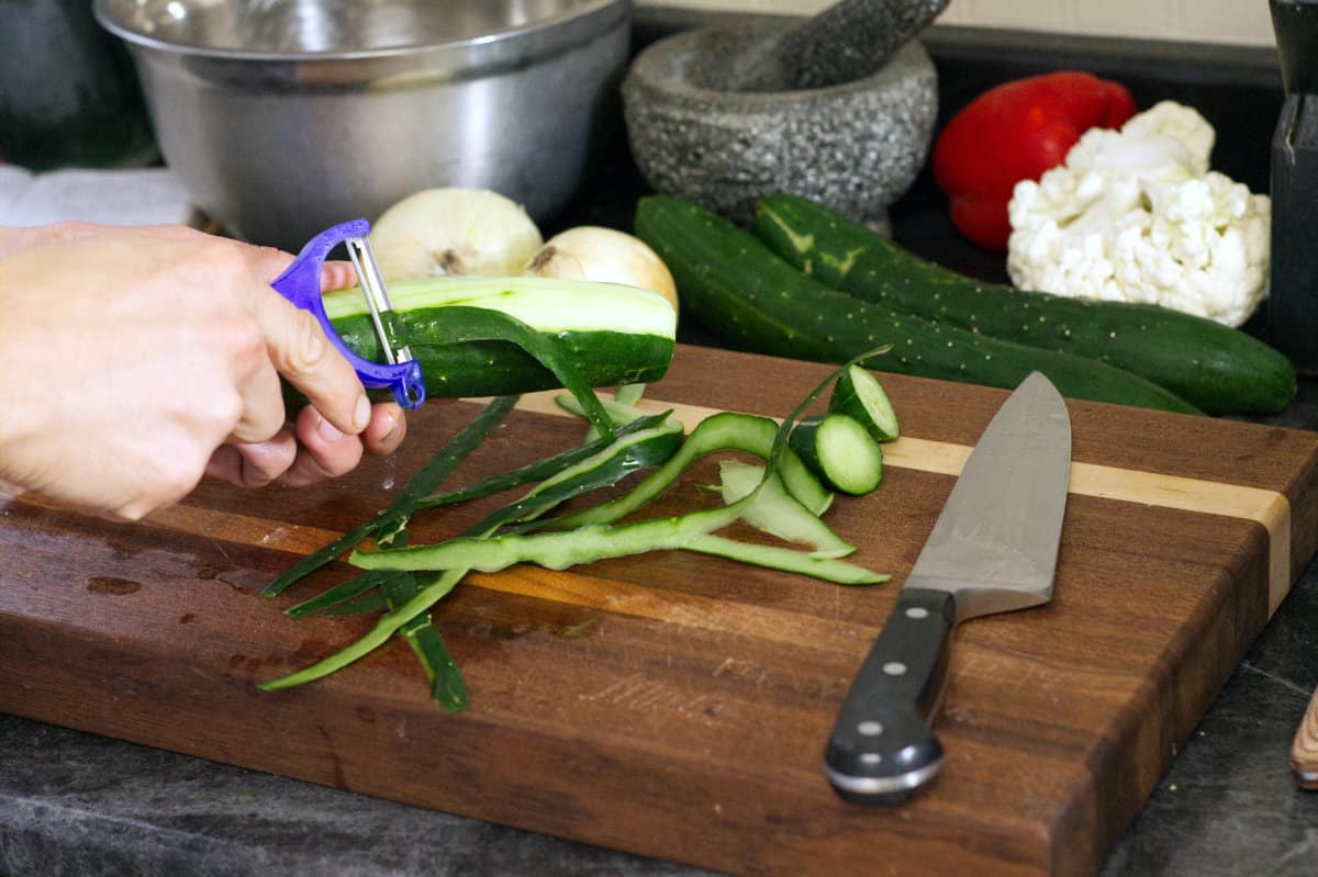 a cucumber being peeled with a blue vegetable peeler. There are various other ingredients in the background.