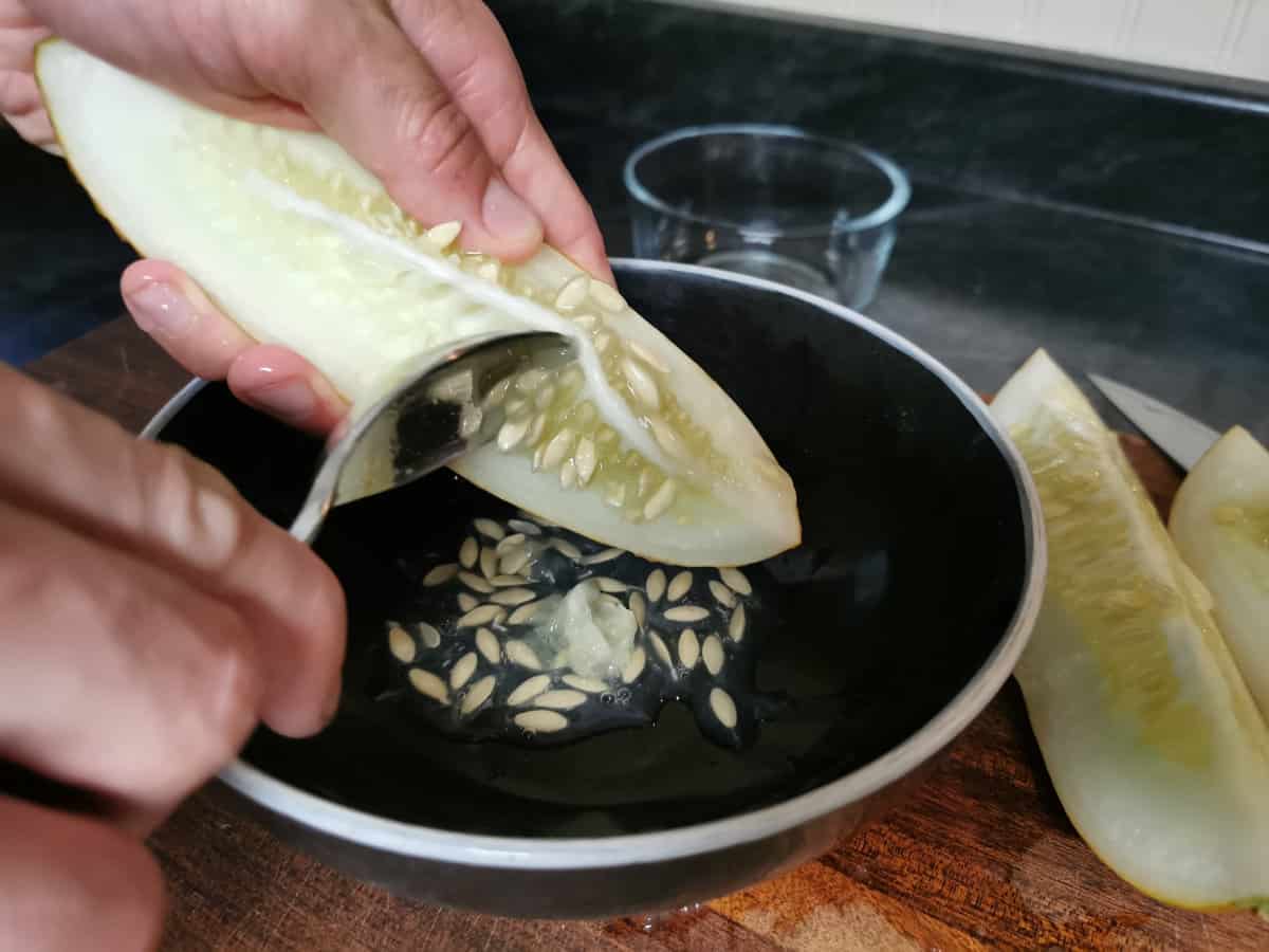 Scooping cucumber seeds out of a quartered cucumber and into a black bowl. The bowl is sitting on a wooden cutting board.