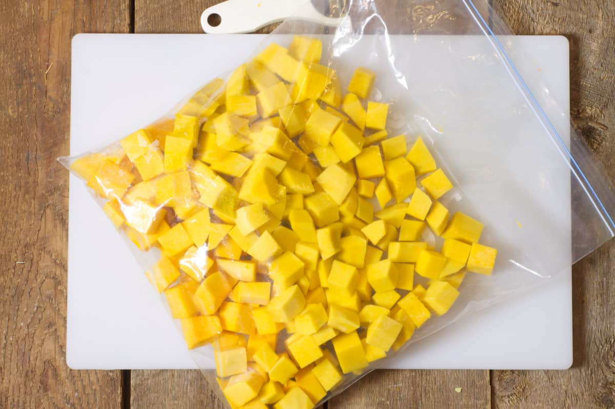 Overhead shot of diced butternut squash in a clear freezer bag laying on a white cutting board. 