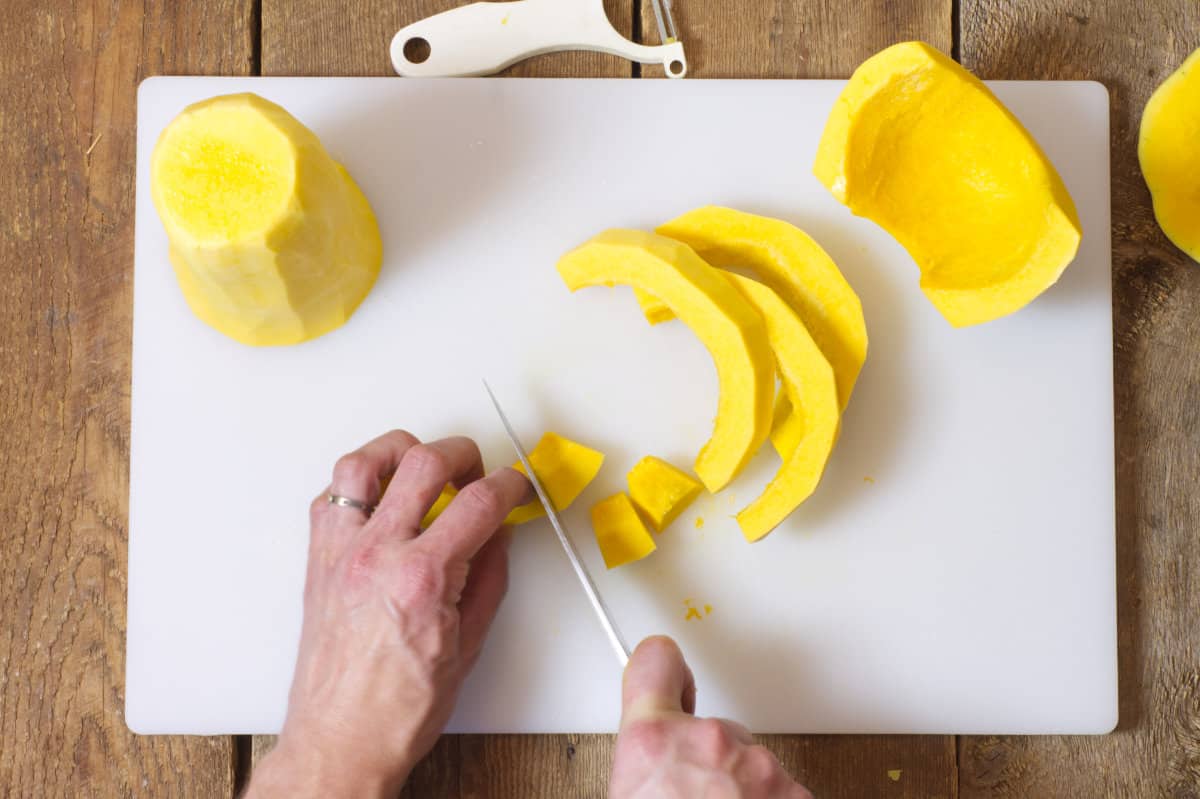 Overhead shot of butternut squash strips being cut into cubes on a white cutting board.