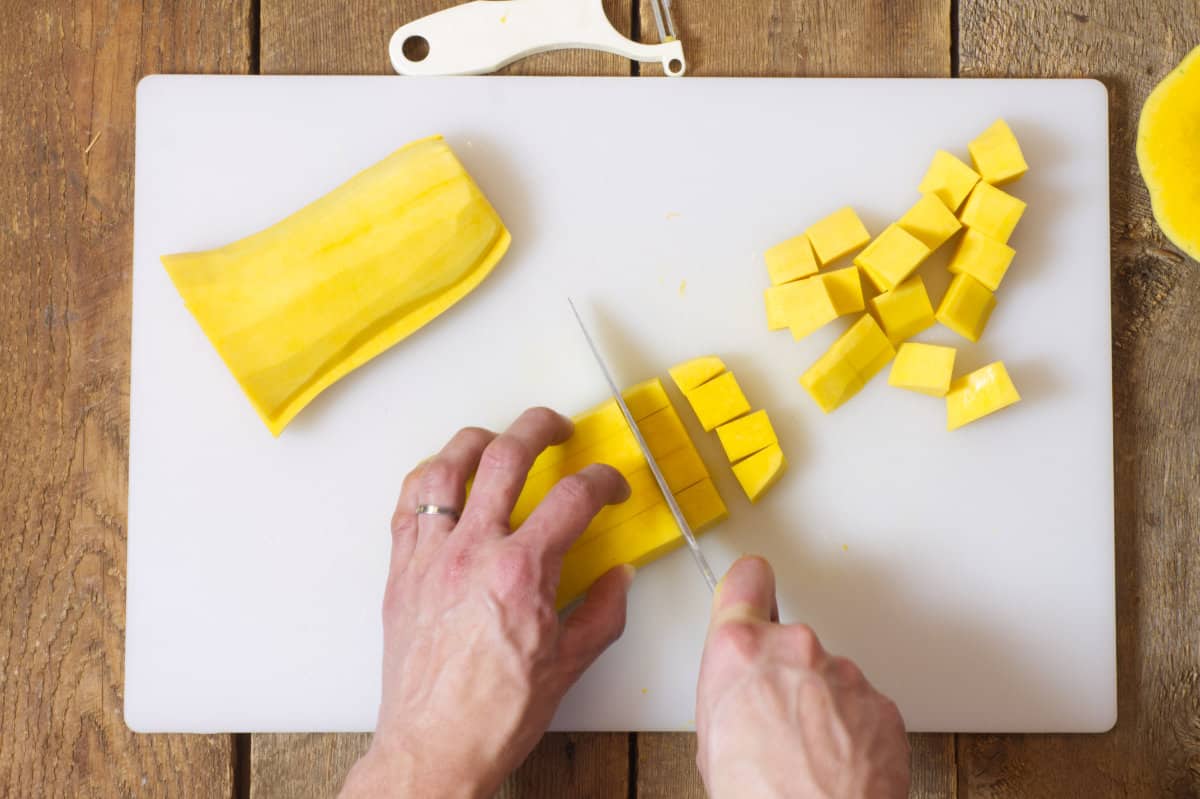 Overhead shot of a peeled butter nit squash being cut into cubes on a white cutting board.