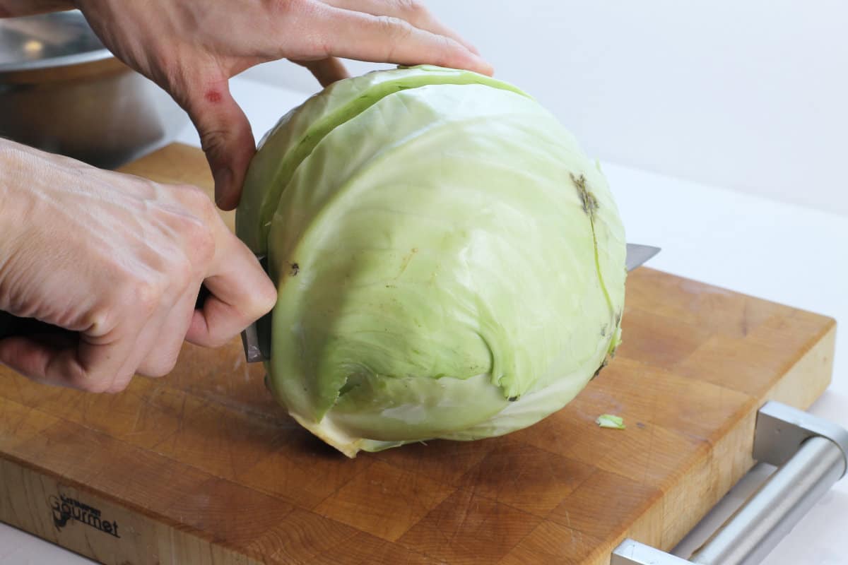 A whole head of cabbage being cut in half with a large chefs knife on a wooden cutting board.