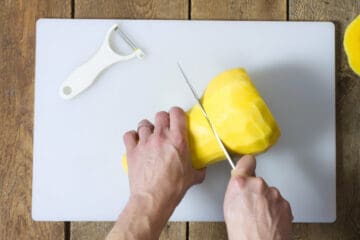 A top down view of a peeled butternut squash being cut in half on a white cutting board.