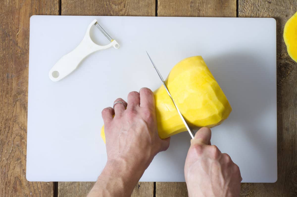 Overhead shot of a peeled butternut squash being cut in half on a white cutting board.