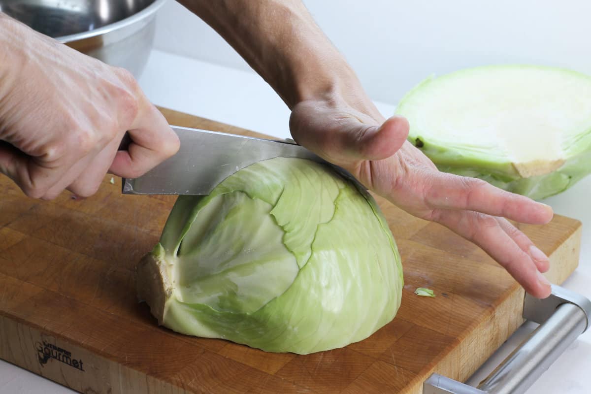 A half of a head of cabbage being cut into quarters. The half head of cabbage is laying flat side down on a wooden cutting board.