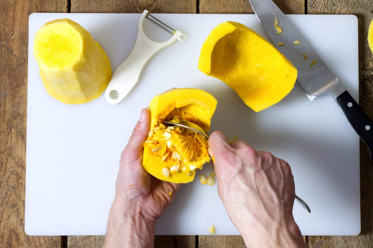 Scooping the seeds out of a cut butternut squash over a white cutting board on a wooden background.