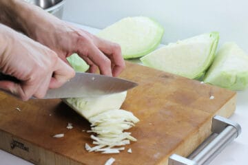 Green cabbage being shredded with a chefs knife on a wooden cutting board.
