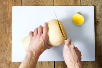 overhead shot of the bottom being cut of a butternut squash on a white cutting board.