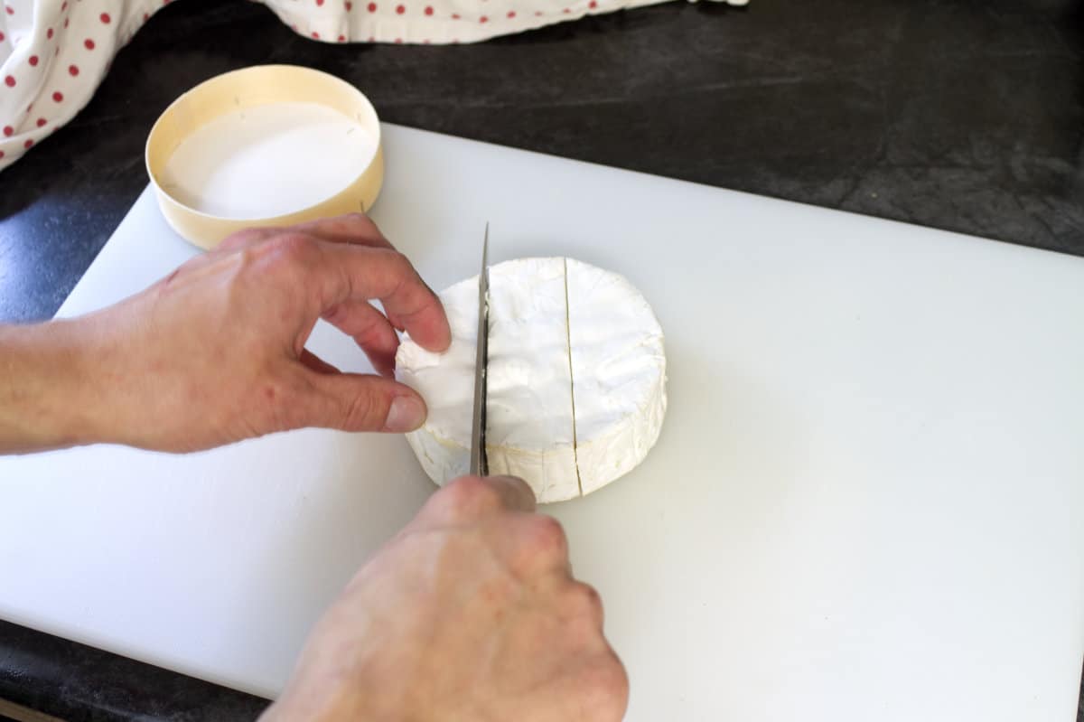A wheel of brie on a white cutting board being cut into thirds.