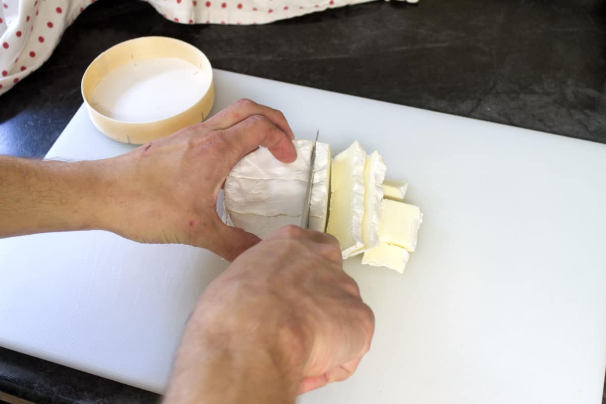A wheel of brie on a white cutting board being cut into thin slices.
