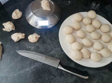 Small balls of bread dough on a white plate besides a scale that is being used to weigh and cut the balls.