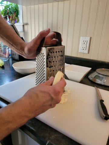 A block of provolone cheese being shredded on a stainless steel box grater.