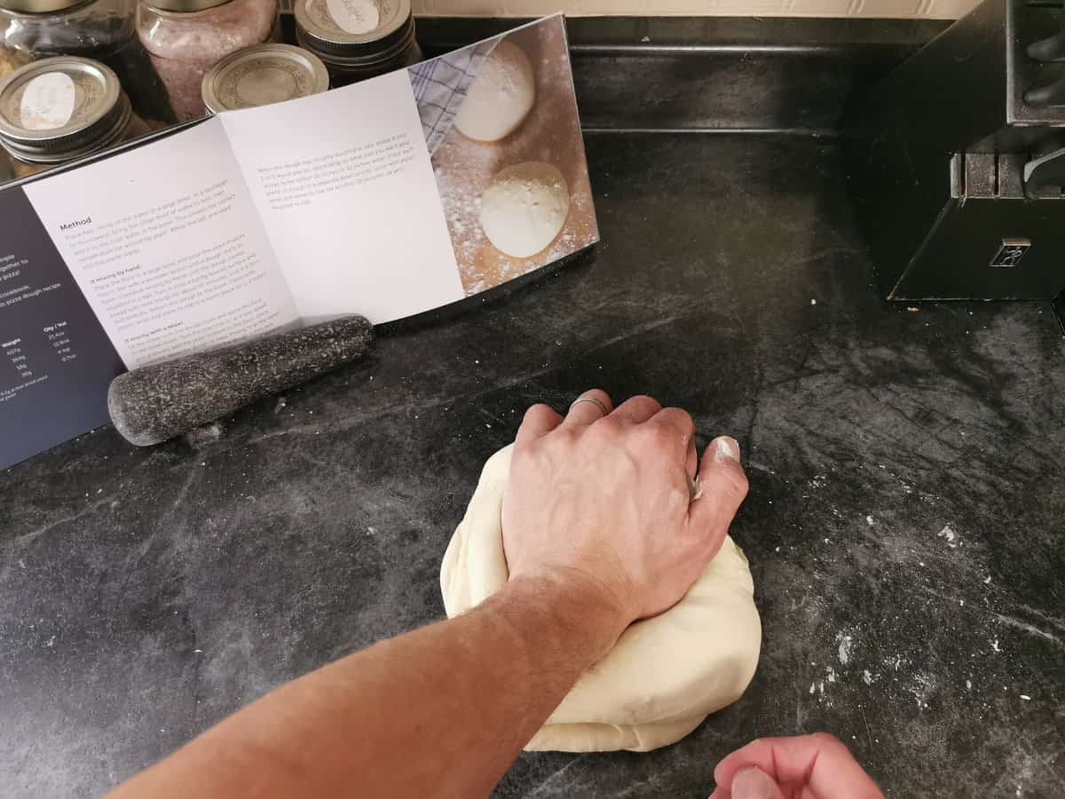 Kneading a ball of pizza dough on a black counter top with a cook book leaned up in the background
