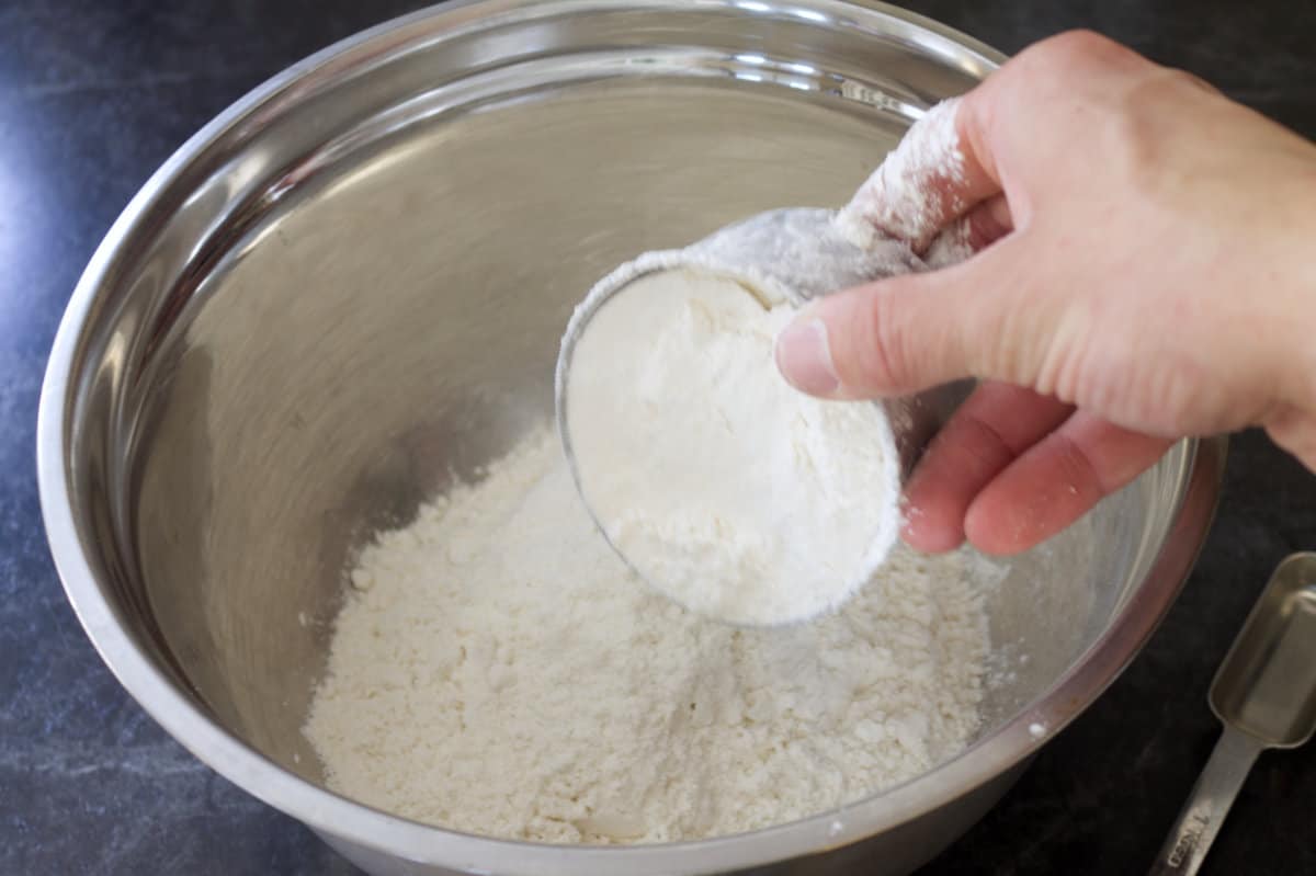 A measuring cup filled with flour being poured into a stainless steel bowl.