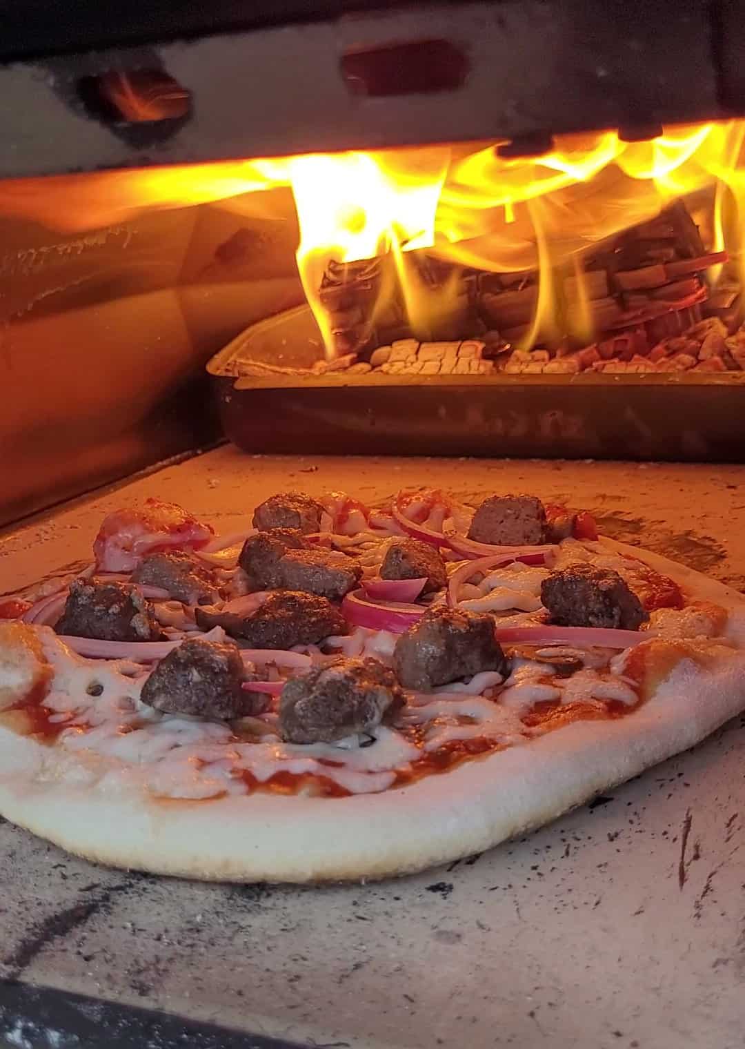 A meatball pizza cooking inside a Ooni pizza oven lit with a wood fire.