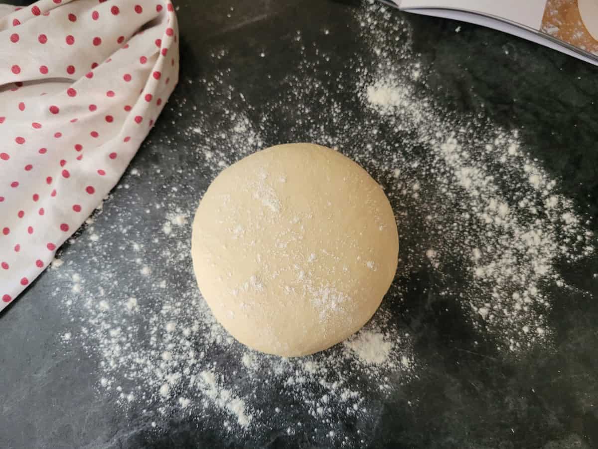 A ball of formed pizza dough, dusted with flour on a black counter top.