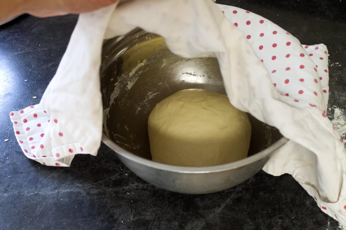 A stainless steel bowl with a ball of dough, partially covered by a red polka dot kitchen towel..