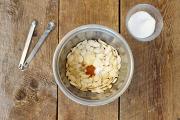 Overhead shot of raw pumpkin seeds in a stainless steel bowl with various spices being mixed in.