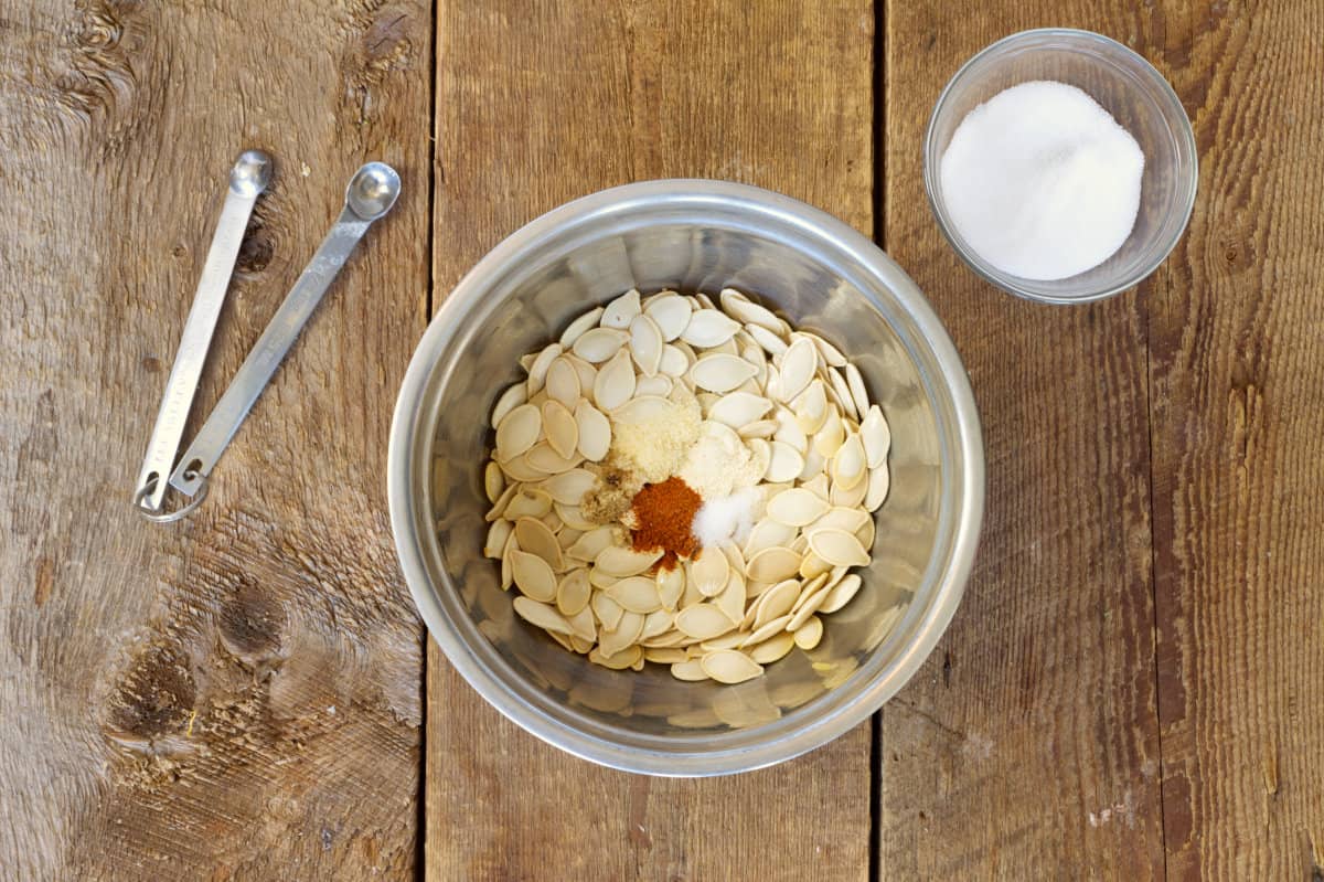 Overhead shot of raw pumpkin seeds in a stainless steel bowl with various spices being mixed in.