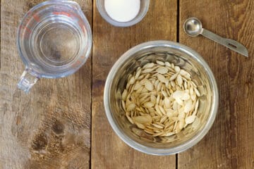 Overhead shot of raw pumpkin seeds in a small stainless steel bowl surrounded by a measuring cup with water, a dish of salt, and a measuring spoon.