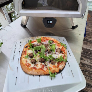 A meatball pizza fully cook and displayed on a perforated steel pizza peel in front of an ooni pizza oven.