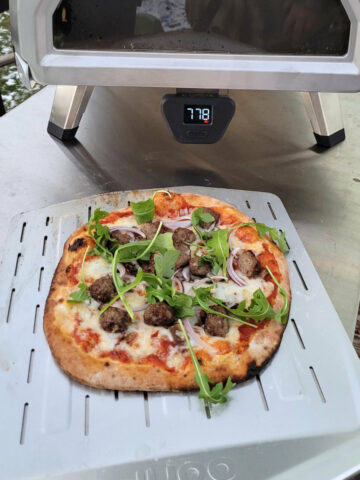A meatball pizza fully cook and displayed on a perforated steel pizza peel in front of an ooni pizza oven.
