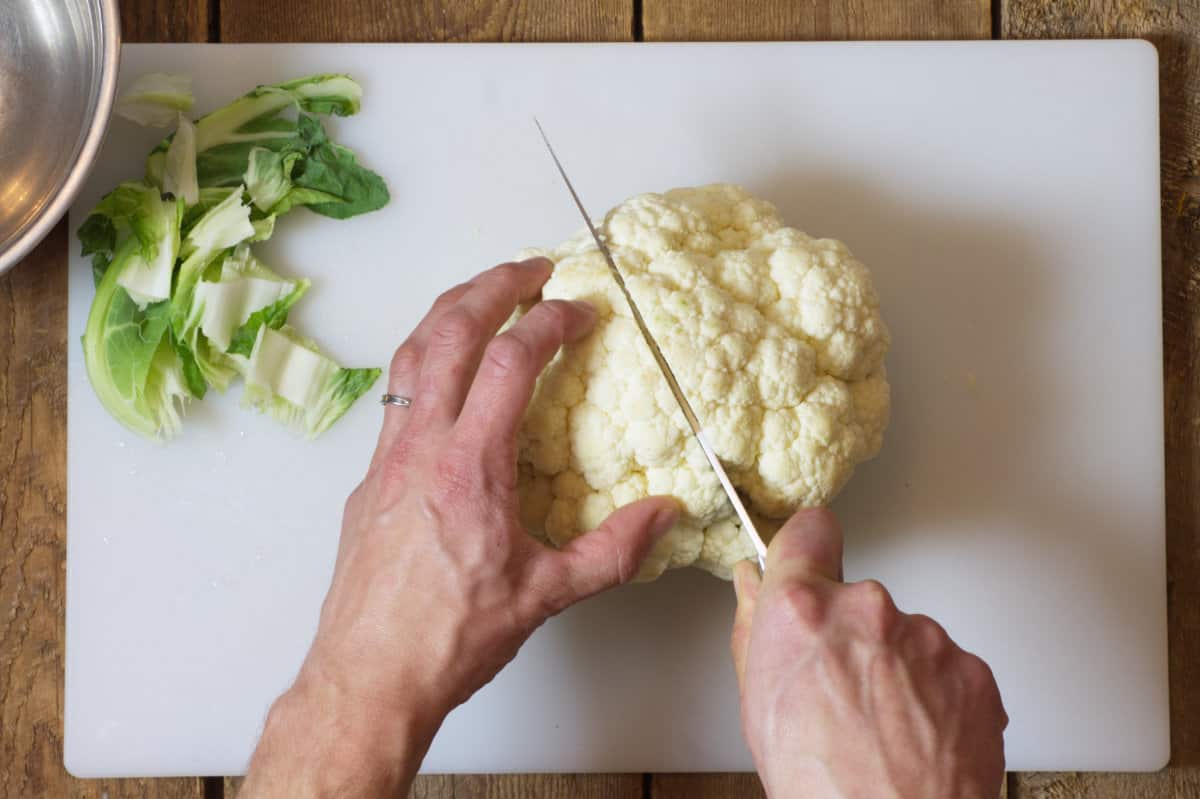 an over head shot of a head of cauliflower being cut in half on a white cutting board.