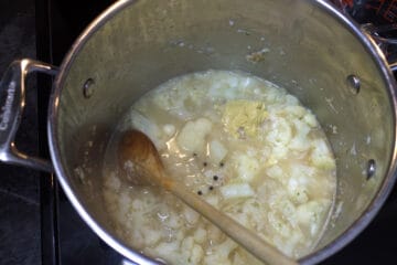 Top down view of a large stainless steel soup pot filled with cooking onion, cauliflower, peppercorns, dijon mustard and chicken stock.