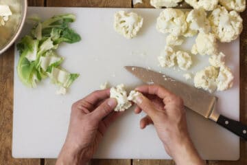 Overhead shot of a white cutting board with cauliflower being broken into small florets by hand.