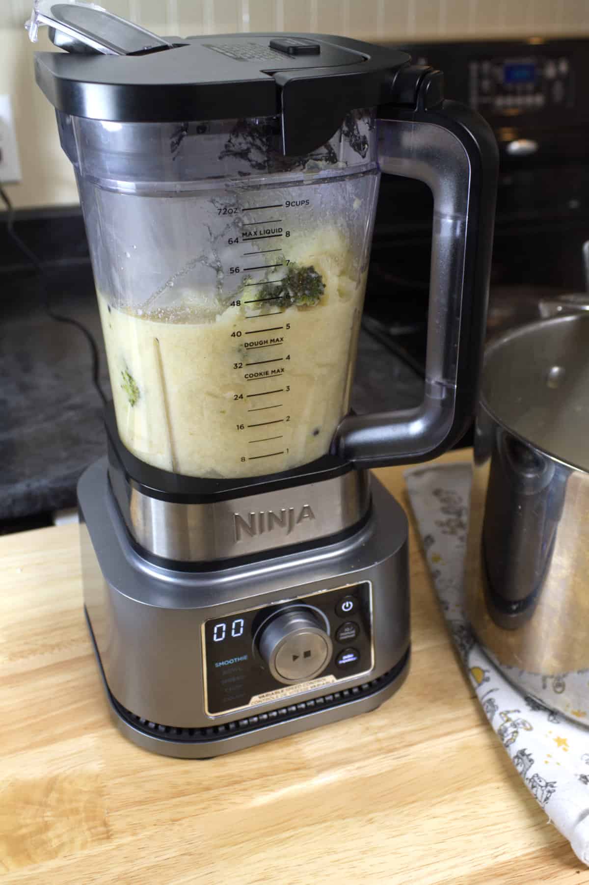 A ninja foodie stand blender on a wood block countertop and filled half full with broccoli and cauliflower soup.