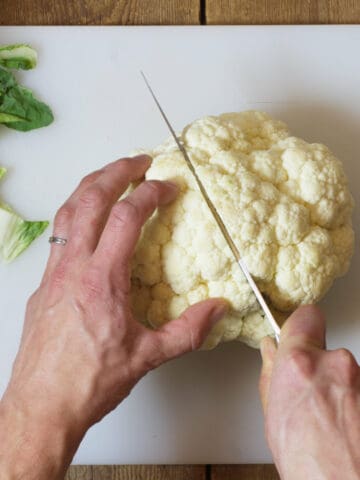 An over head shot of a head of cauliflower being cut in half on a white cutting board.