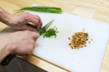 Chives being finely chopped on a white cutting board beside a small pile of chopped roasted almonds.