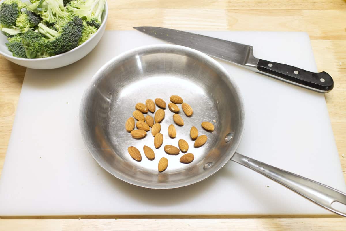 Whole almonds in a steel pan which is sitting on a white cutting board, surrounded by a bowl of broccoli and a chefs knife.