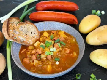 Bowl of potato goulash with toasted bread sitting on the table, with basic ingredients - potato and sausage - sitting next to the goulash.