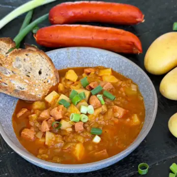 Bowl of potato goulash with toasted bread sitting on the table, with basic ingredients - potato and sausage - sitting next to the goulash.