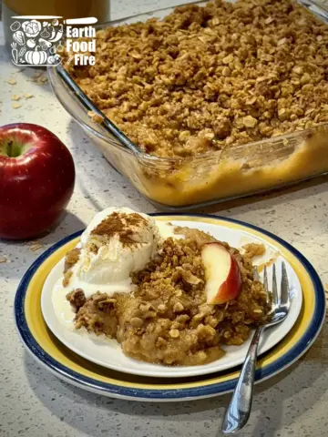 An applesauce crisp served with ice cream on a plate, with full baking tray of applesauce crisp in the background