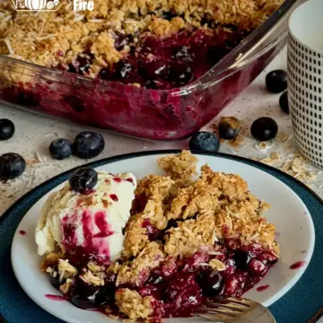 The results of the blueberry crumble recipe: blueberry crumble served with ice cream on a plate with the full baking tray of the blueberry crumble in background