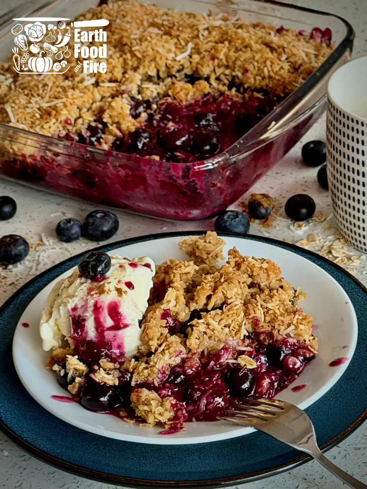 The results of the blueberry crumble recipe: blueberry crumble served with ice cream on a plate with the full baking tray of the blueberry crumble in background