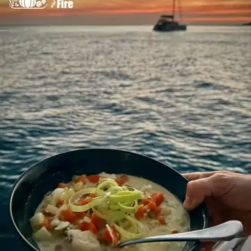 A steaming bowl of cod fish chowder with chunks of white fish, potato, carrot, and leek in a creamy broth, garnished with fresh leek.