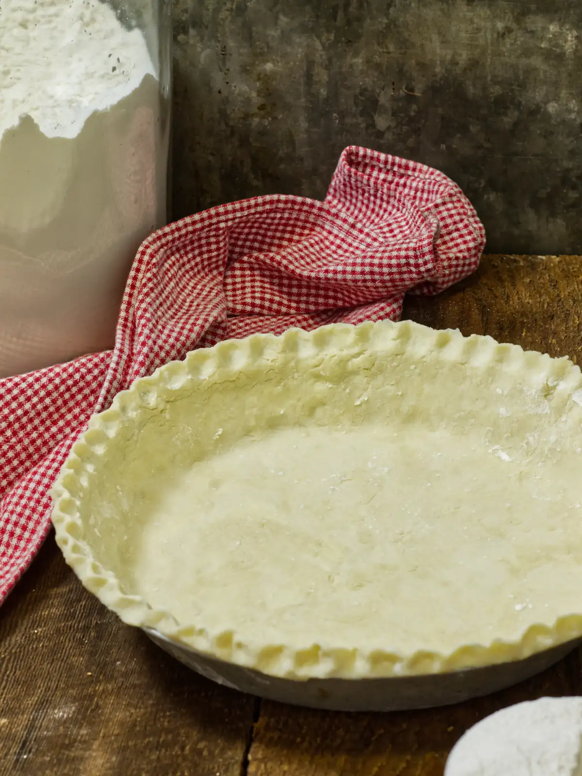 Finished result of our homemade pie shell recipe sitting on a wooden table, with a jar of flour and kitchen towel