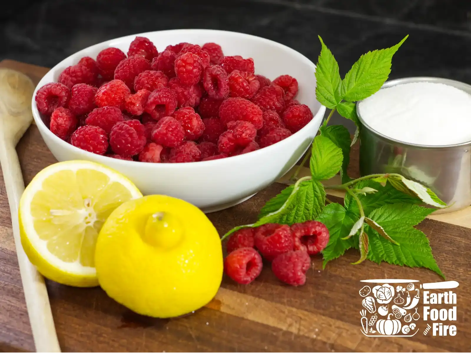 Raspberry cordial ingredients laid out on a wooden table - fresh raspberries, a halved lemon, and a bowl of white sugar.