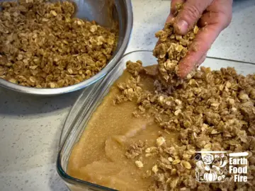 The crumble topping being scattered by hand over the applesauce filling